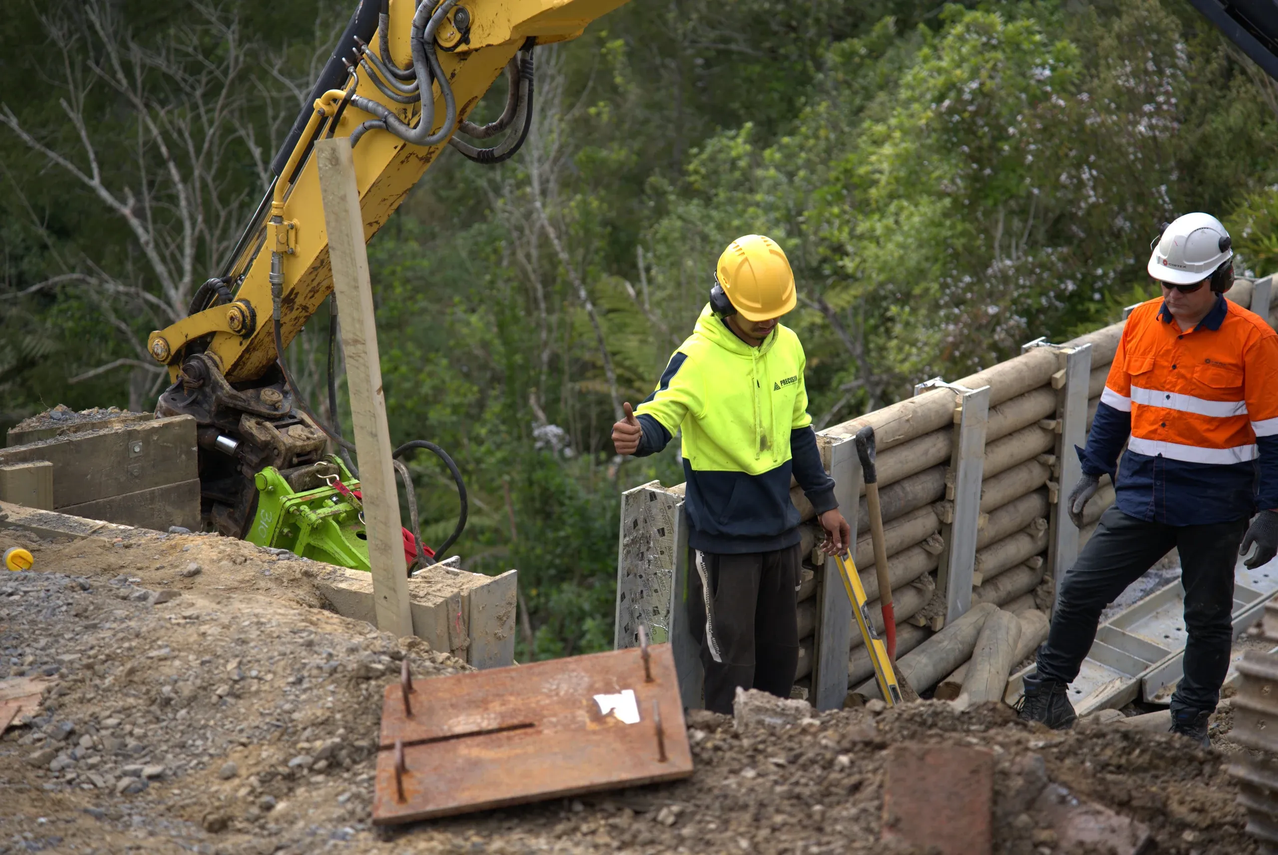 Crew setting out timber lagging between the installed soldier piles while the excavator holds position above the wall.