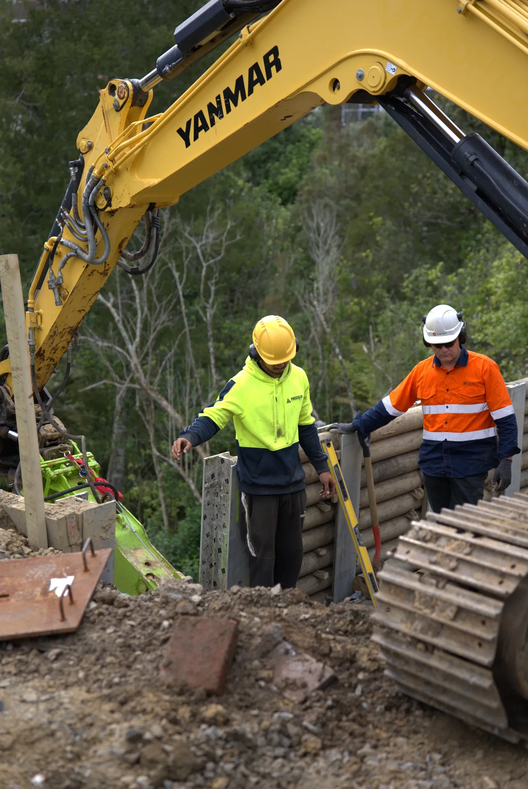 Excavator and crew lowering timber lagging boards into place along the pile line.