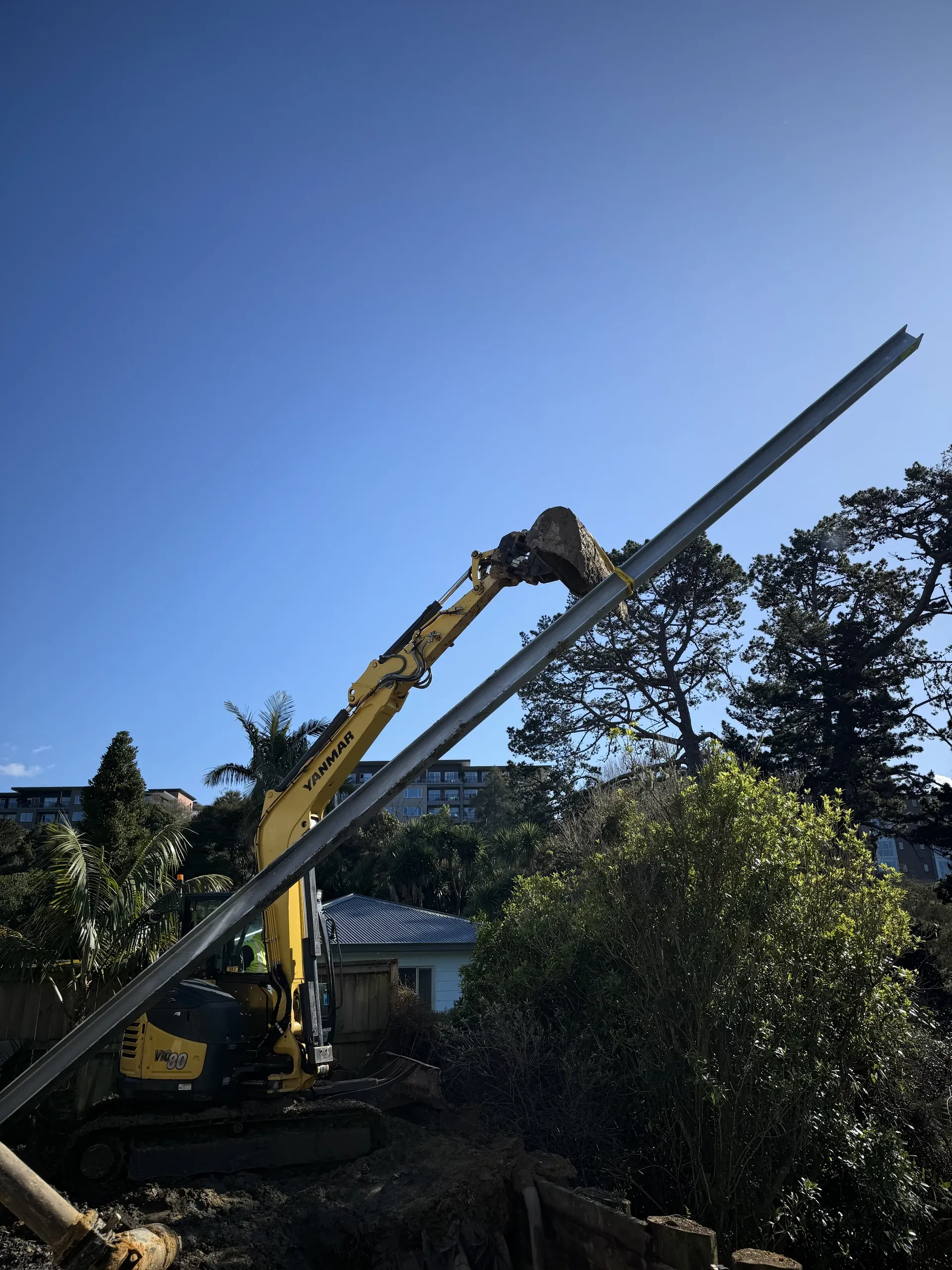 Excavator lifting a steel I-beam soldier pile upright against a clear sky before installation.