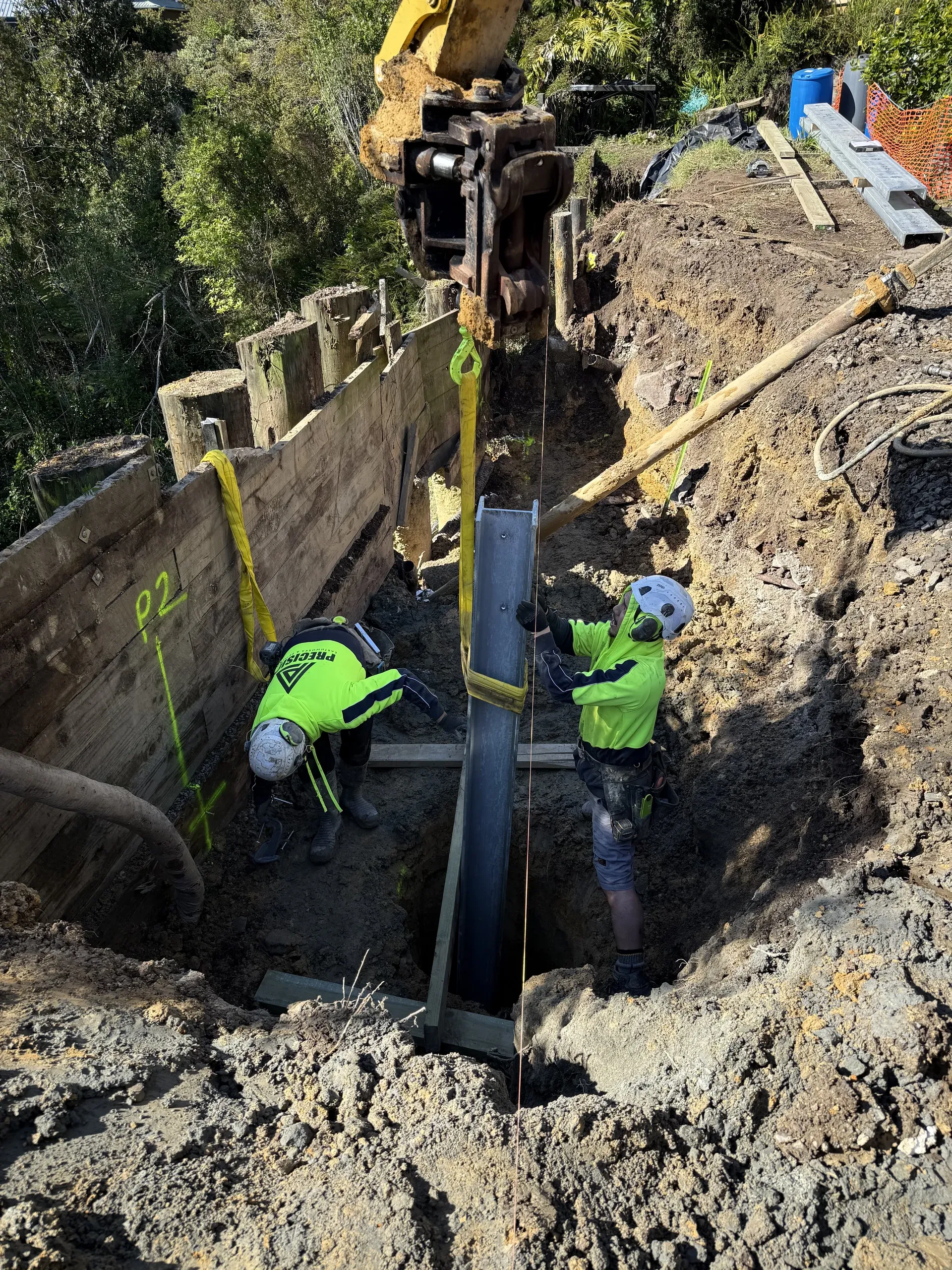 Two crew guiding a steel I-beam soldier pile down into its bored hole alongside the timber-lagged wall.