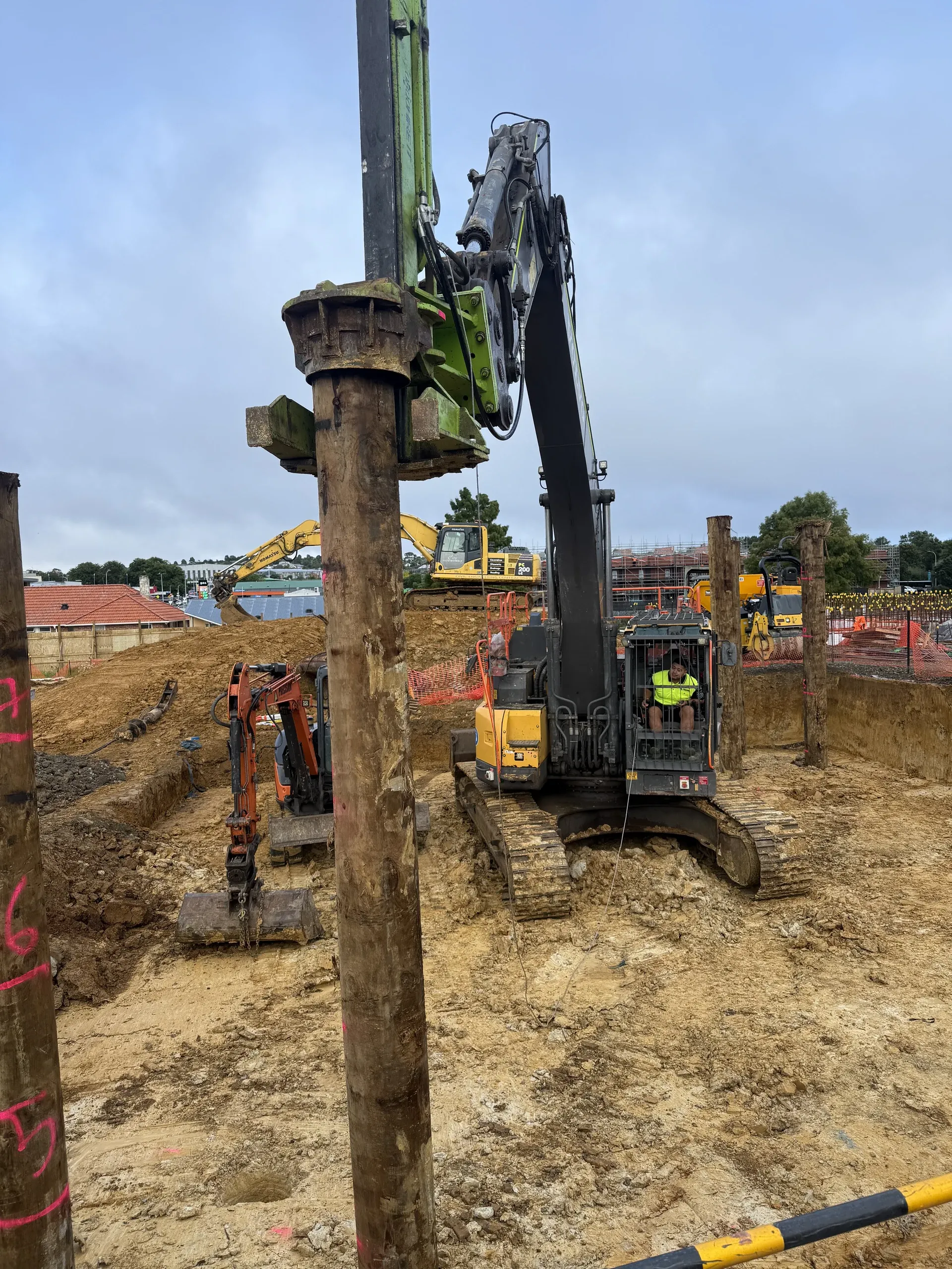 Piling head clamped over a pile as it is driven, with numbered piles already installed across the excavated platform.