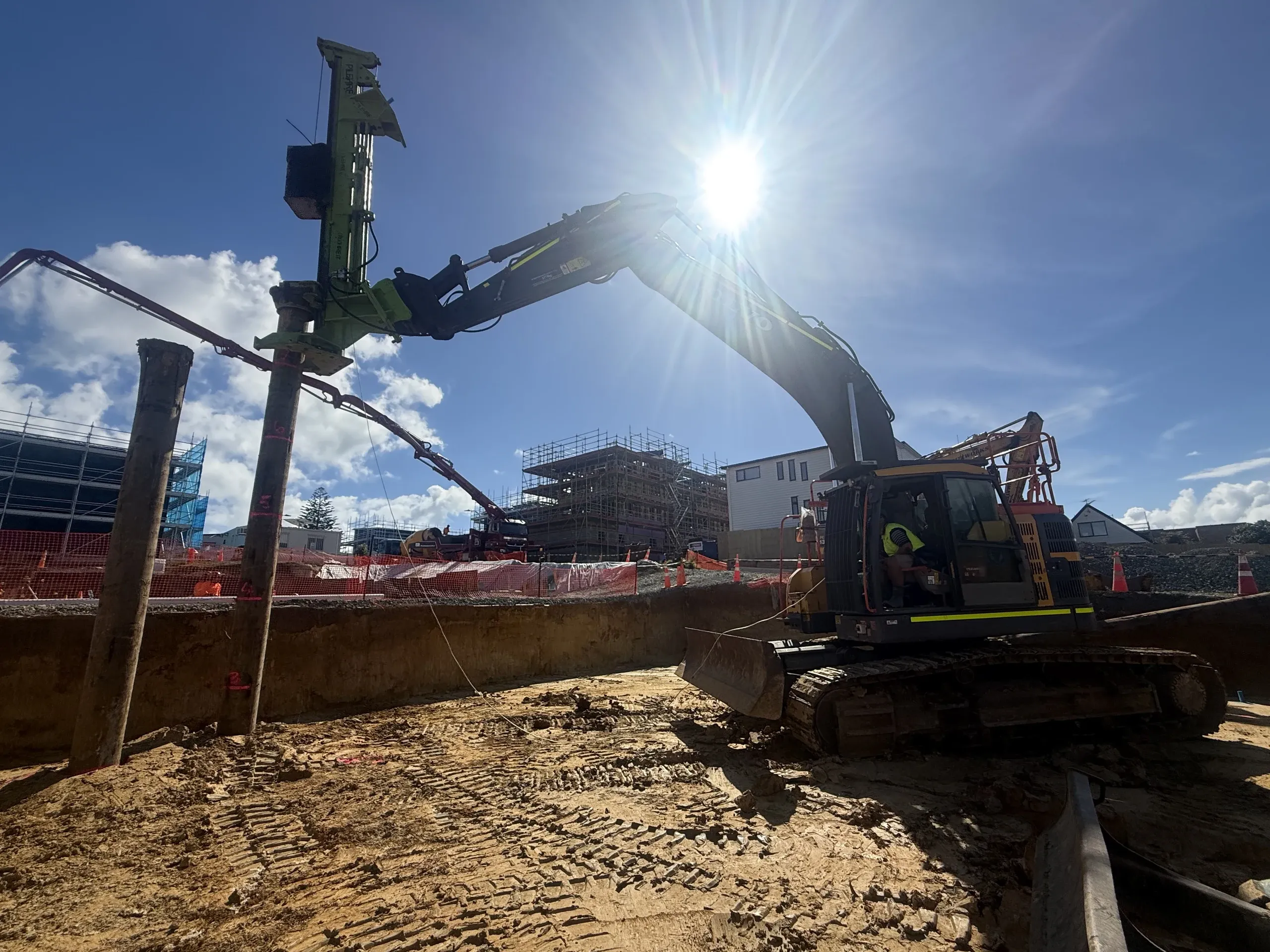 Excavator-mounted piling rig driving a pile on the cut platform, with the subdivision's scaffolded builds rising behind.