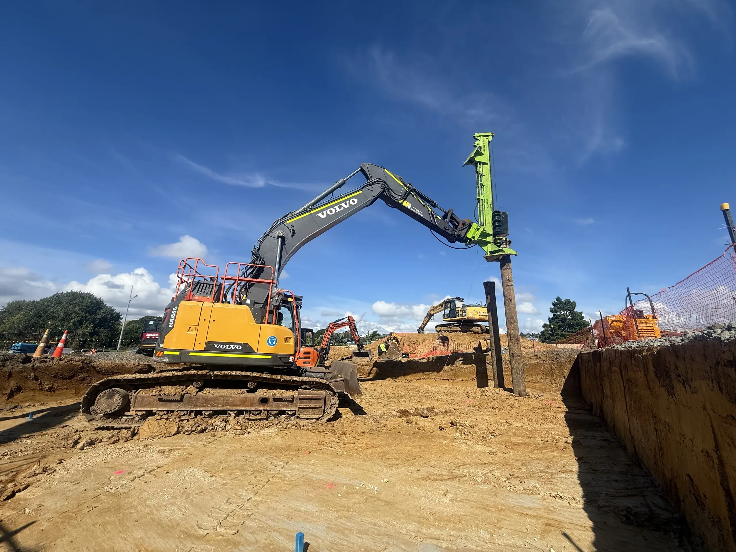 Excavator working alongside installed posts, showing the scale of the retaining run across the site.